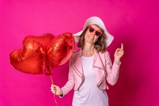 A Portrait of a Caucasian woman having fun dancing with a white hat and some heart balloons on isolated pink background