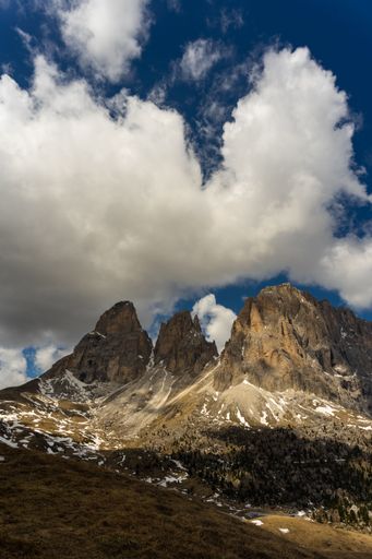 A vertical shot of the Dolomite Mountains with thick clouds above during daylight with sunrays and shadows on it