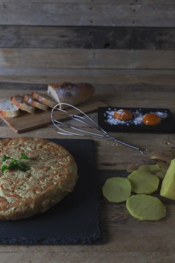 A vertical shot of a potato omelet on a slate board surrounded with pre-preparation ingredients