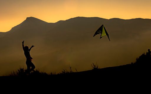 The silhouette of a kid flying a kite surrounded by mountains during the sunset