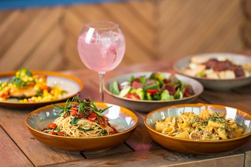 A closeup of pasta and salads with vegetables and meat slices on plates with a glass of cocktail
