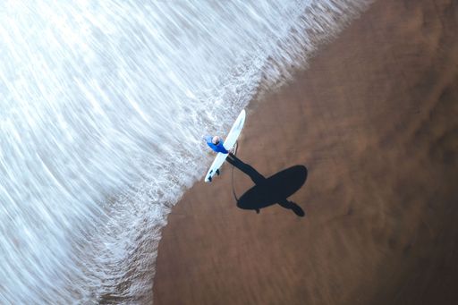 An aerial top view of a surfer walking on a sandy beach against a sea, holding a surf board in Costa Rica shot in long exposure