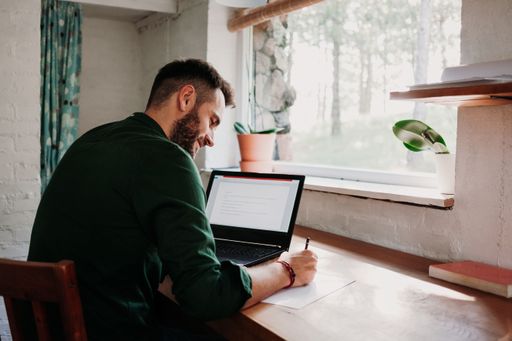 A young Caucasian male taking notes while working on a laptop at home