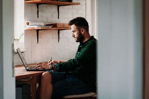 A young Caucasian male taking notes while working on a laptop at home