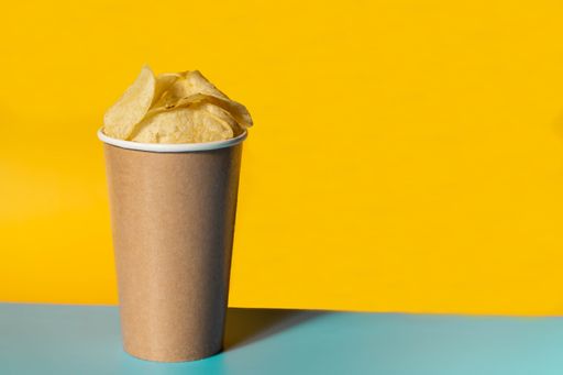 A closeup shot of plain potato chips in a beige paper cup on a yellow background