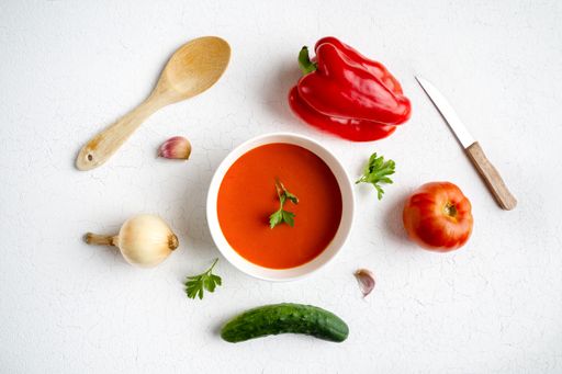 An overhead shot of gazpacho soup surrounded with bell pepper, tomato, garlic, cucumber, onions, parsley and a wooden spoon and knife on a white table
