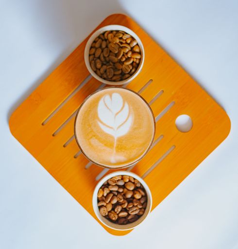 A vertical closeup of the cup of Cappuccino with coffee beans on the orange board on a white table.