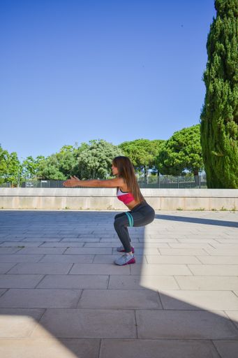 An athletic young Caucasian female working out and doing squats with elastic  resistance bands in a park