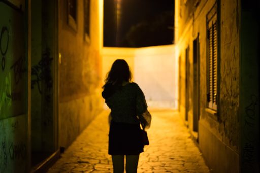A female with long hair walking through a dimly lit alley at night shot from behind