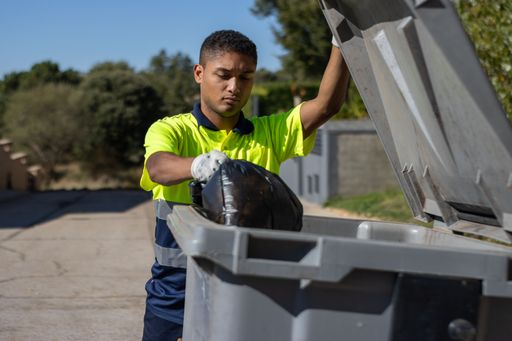 A worker throws a bag of garbage into a large dumpster while holding up the dumpster lid