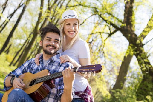 A happy caucasian couple with the man playing guitar while sitting on a bench, his lover behind him