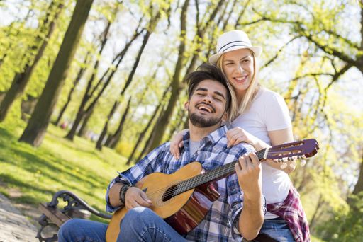 A happy caucasian couple with the man playing guitar while sitting on a bench, his lover behind him