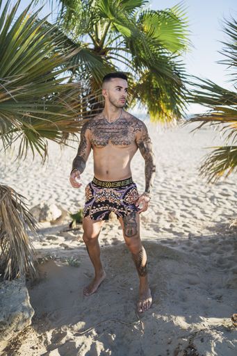 A vertical shot of a Caucasian male with tattoos on his chest, arms, and legs wearing fashionable clothes posing at the beach