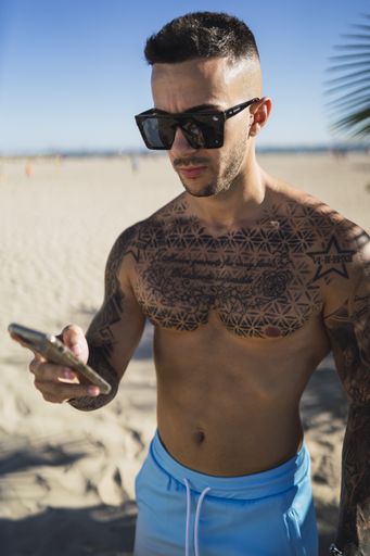 A vertical shot of a Caucasian male with tattoos wearing sunglasses while using a smartphone at the beach