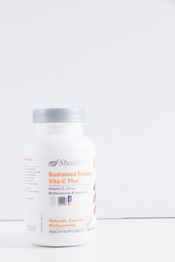 A vertical closeup of a white plastic bottle of Vitamin C "Sustained Release Vita-C Plus" against a white wall