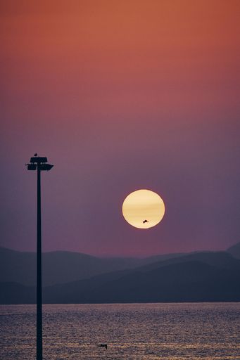 A silhouette of a lamp post near the sea with the sun going down behind the mountains at sunset