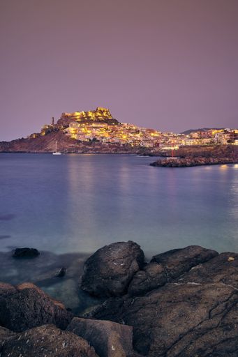 A vertical shot of buildings with lit lights near the sea and a purple sky in the background at sunset