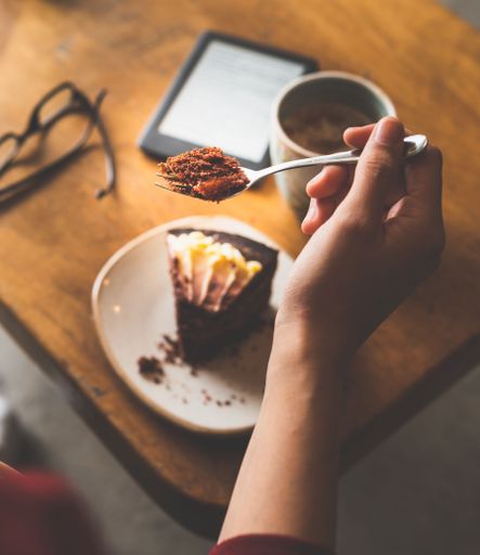 A vertical shot of a hand picking up a piece of cake from a cafe table with coffee and an e-reader