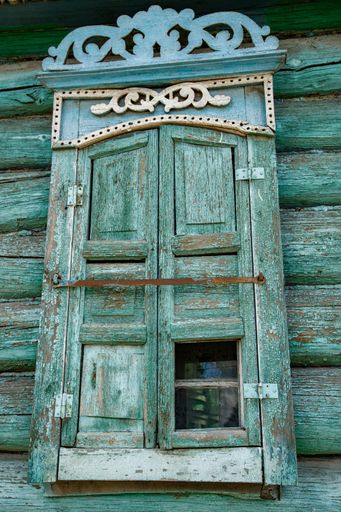 A vertical shot of an old dilapidated log house window with carved platbands and shutters