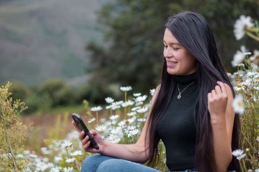A closeup shot of a cheerful woman sitting on the field surrounded by white flowers and looking at her phone