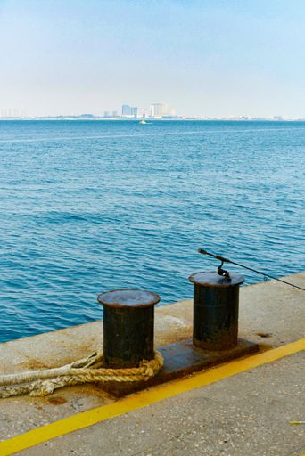 A vertical shot of a pier with mooring bollards, overlooking a turquoise sea and buildings on the horizon
