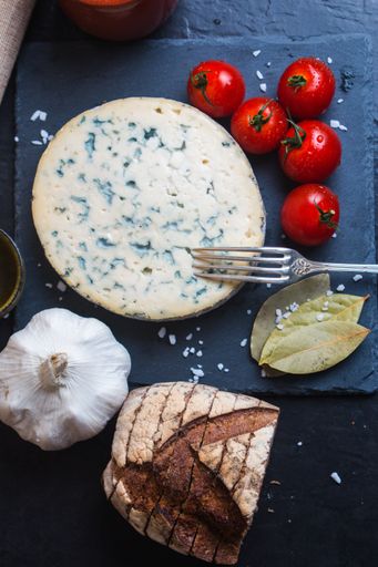 A vertical shot of the blue cheese next to fork, tomatoes, garlic, bread and bay leaf