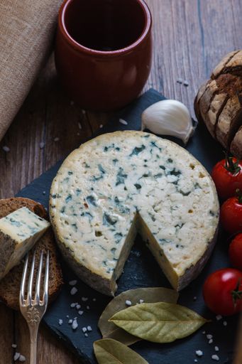 A vertical shot of the blue cheese next to fork, cup, tomatoes, garlic, bread and bay leaves