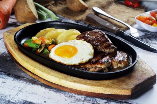 A closeup of a typical Indonesian steak served with vegetables and fried egg in a metal tray on a wooden table