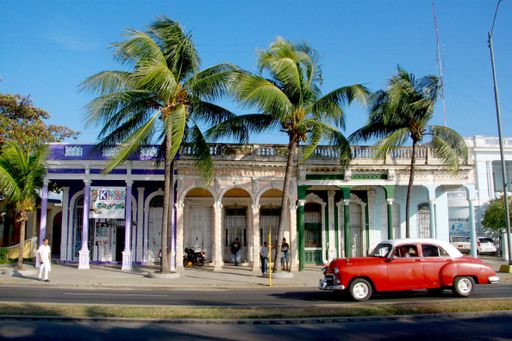 An old red car on the side of the road in front of a building with palm trees in Malecon de Cienfuegos