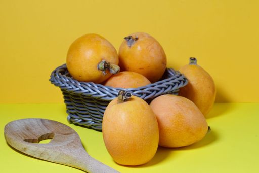 A closeup of medlars in a small wicker basket and a wooden spatula on yellow background