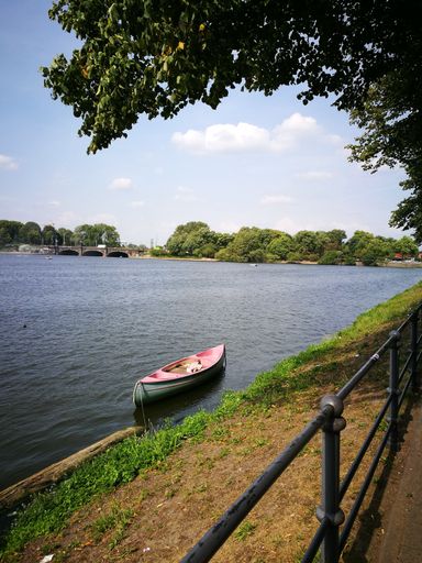 A vertical shot of a small pink boat in the side of a river near a park with no people during midday
