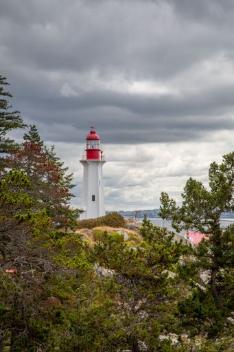 A vertical shot of a lighthouse by the coast in the background of a lake full of sailboats in the cloudy weather.