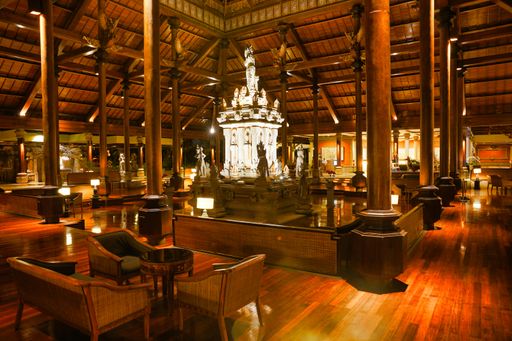 A beautiful statue in the center of a resort lobby with brown chairs and tables under a wooden roof