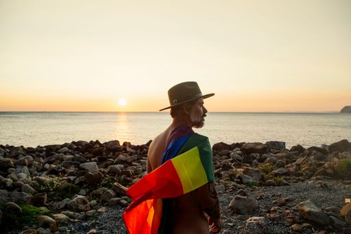 The man with an LGBT flag standing on the shore at sunset.