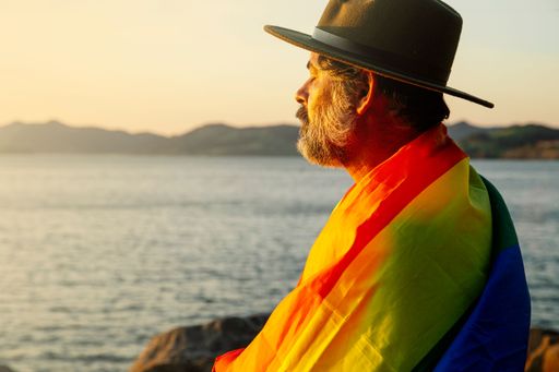 A closeup of a man with an LGBT flag sitting on the shore under the light of sunrise.