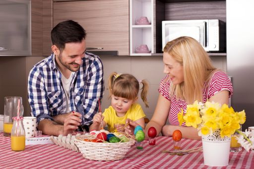 A caucasian family sitting behind the kitchen counter with their daughter and painting Easter eggs