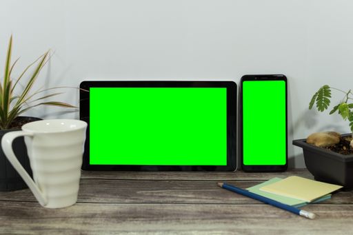 A tablet and a phone with green screens placed on a table with a cup in front of it
