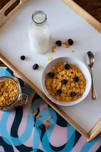 A bowl of corn flakes with blackberries next to the milk and corn flake jars