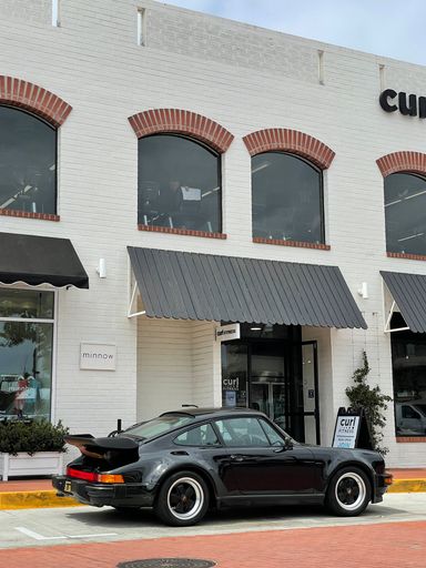 A vertical shot of a black classic Porsche Turbo 930 parked near the white building in Newport Beach