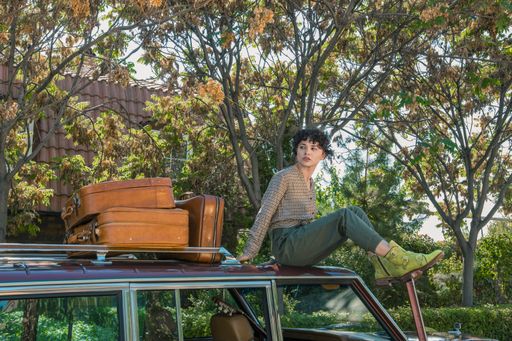A female model sitting on a car posing for a photoshoot with suticases to the side on a bright sunny day