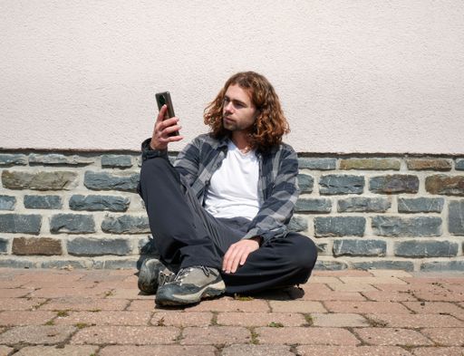 A Caucasian guy sitting on the ground outdoors, using his phone