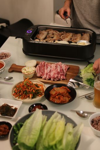 A vertical shot of a dining table with an indoor grill, meat on a board, plates of vegetables