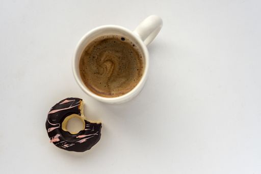 An overhead shot of a tasty doughnut with a cup of coffee on a white-colored surface