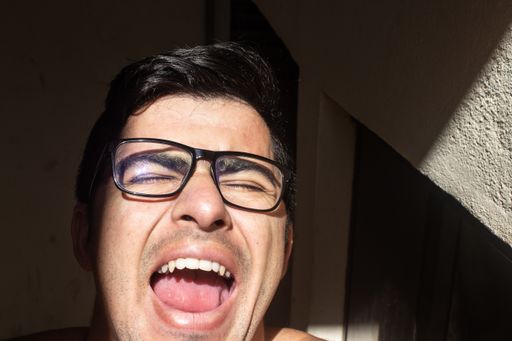 A closeup portrait of a young Hispanic man wearing eyeglasses screaming in the balcony with sunlight on his face