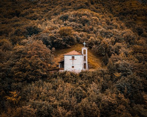 A bird's-eye view of a church standing on top of fall colored hill ,surrounded by a forest at sunset in Dolomites.