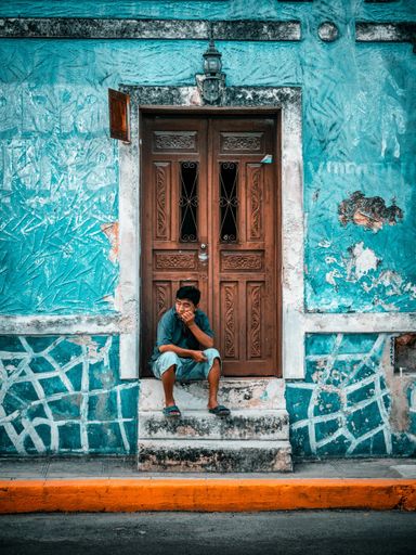 A vertical shot of a man sitting on the stairs in front of his door. An old turquoise building with a brown ornamental door