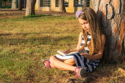 The little girl reading a book while sitting under a tree in the park.