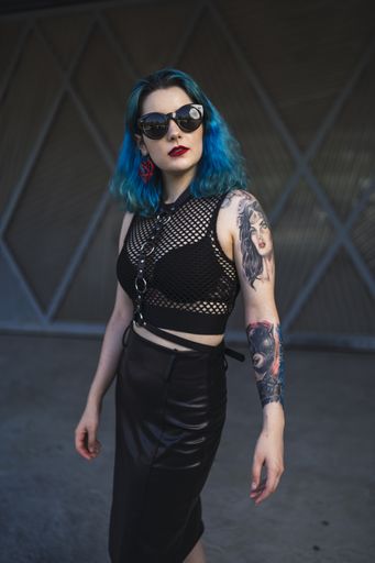 A European female with tattoo and blue hair wearing sunglasses and sexy black dress posing at the side of a building