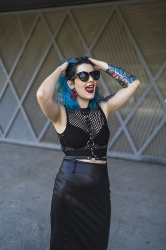 A European female with tattoo and blue hair wearing sunglasses and sexy black dress posing at the side of a building