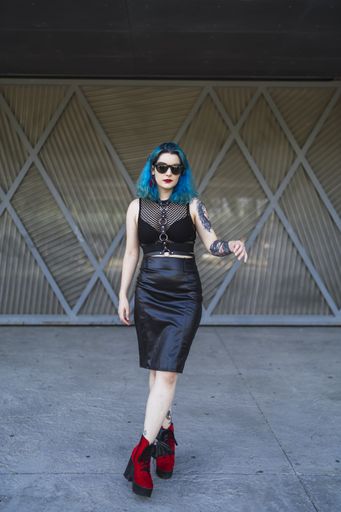 A cool young lady with blue hair, tattoos, and sunglasses, wearing a crop top, leather pencil skirt and red shoes posing in front of a garage